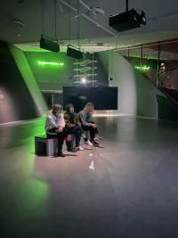 In an indoor space, three people sit on a bench, facing the same direction. Behind them, there is a large screen, seen from the back, and a sentence in green neon lights, which hangs on a wall.