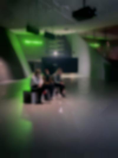 In an indoor space, three people sit on a bench, facing the same direction. Behind them, there is a large screen, seen from the back, and a sentence in green neon lights, which hangs on a wall.