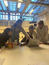 Four people sit or stand next to a table. One of the people is pointing at the screen of a grey laptop, which is open on the table. There is a cup, an open bag of crisps and a sheet of paper also on the table.