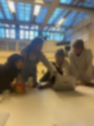 Four people sit or stand next to a table. One of the people is pointing at the screen of a grey laptop, which is open on the table. There is a cup, an open bag of crisps and a sheet of paper also on the table.