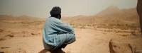 A person, squatting down, faces an arid area, with hills at the distance. They appear to be wearing a light blue long garment and a turban.