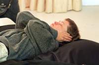 A person is lying supine on a large black cushion. They have short hair and are wearing a grey sweater. Their hands are covering their ears and their eyes are closed.