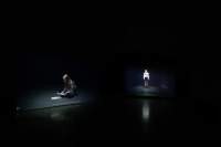 Two people appear, as separate images, against a black background. They both appear to be on stage, with a spotlight on them. On the left, a young person with long hair sits on their heels, reading a piece of paper. More paper lies on the floor in front of them. On the right, the other person is at the distance, standing up straight.