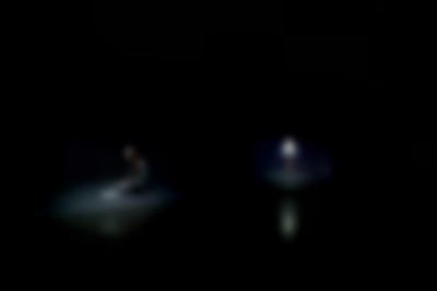 Two people appear, as separate images, against a black background. They both appear to be on stage, with a spotlight on them. On the left, a young person with long hair sits on their heels, reading a piece of paper. More paper lies on the floor in front of them. On the right, the other person is at the distance, standing up straight.