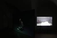Two images appear, separately, against a black background. On the left, in high transparency, a young person with long hair sits on their heels, reading a piece of paper. More paper lies on the floor in front of them. On the right, a sheet of paper appears in front of the camera. Behind it, there is a microphone stand and a few people.