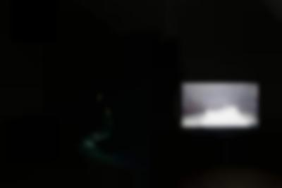 Two images appear, separately, against a black background. On the left, in high transparency, a young person with long hair sits on their heels, reading a piece of paper. More paper lies on the floor in front of them. On the right, a sheet of paper appears in front of the camera. Behind it, there is a microphone stand and a few people.