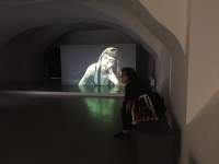 At the end of a room, a large screen shows the image of a person with a short fringe and long straight hair. Their head is tilted to the left, resting on their left fingers. In the room, a person sits near one of the walls. They are facing a different direction than that of the screen.