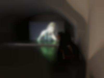 At the end of a room, a large screen shows the image of a person with a short fringe and long straight hair. Their head is tilted to the left, resting on their left fingers. In the room, a person sits near one of the walls. They are facing a different direction than that of the screen.