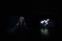 Two people appear, as separate images, against a black background. One is an older child with long hair, facing the camera, and the other a person leaning back. They are only visible from the torso up.