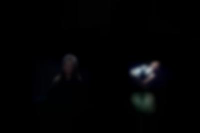 Two people appear, as separate images, against a black background. One is an older child with long hair, facing the camera, and the other a person leaning back. They are only visible from the torso up.