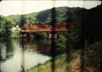River with red bridge. There is green vegetation on the river banks. Several bars are out of focus, in the forefront of the photograph.
