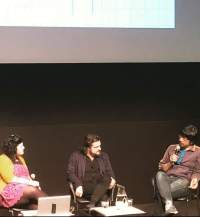 Three people sit under a screen. The person on the right is holding a microphone to their mouth. The other two people look towards them. There is an open laptop in front of the person on the left, and a low table with a water jug and glasses in front of the person in the middle.