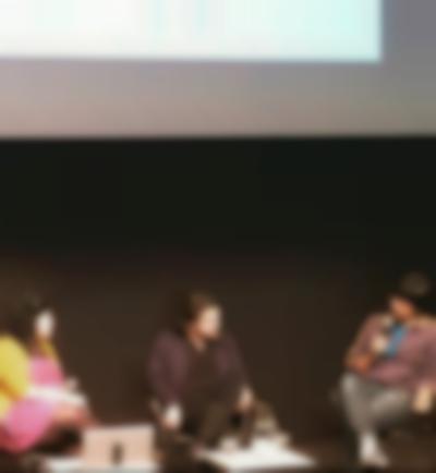 Three people sit under a screen. The person on the right is holding a microphone to their mouth. The other two people look towards them. There is an open laptop in front of the person on the left, and a low table with a water jug and glasses in front of the person in the middle.