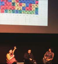 Three people are sitting underneath the projection of an image. The person on the left is holding the microphone and gesturing towards the screen with their left hand. The image shows square paper with squares hand-painted in different colours. Each square has a number inside.