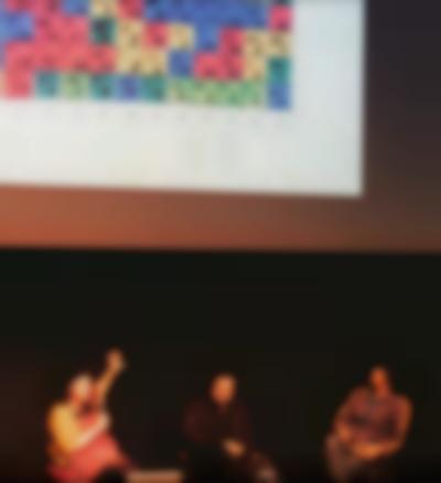 Three people are sitting underneath the projection of an image. The person on the left is holding the microphone and gesturing towards the screen with their left hand. The image shows square paper with squares hand-painted in different colours. Each square has a number inside.