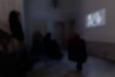 Three people watch a film being projected on a wall of a hallway space. Two of them are sitting down, while the other stands up at the bottom of the stairs.