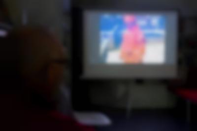 A person looks towards a pull down projector, where a film is being projected. The image shows a person in a full orange suit and hat.
