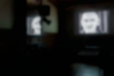A film is being projected on a screen. The image shows a head shot of a masculine-presenting person with a moustache. The image also appears partly reflected on a mirror to the left.