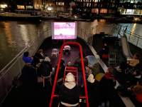 On a deck, viewed from above, a group of people stand or sit in front of a screen where something is being projected. To their right, there is a slide. In front of the screen, there is a red playground overhead ladder, behind which someone is standing, wearing headphones. In the background, behind the screen, there is a river and two large buildings on the other bank. It is dark and several of the windows in the buildings have lights on.