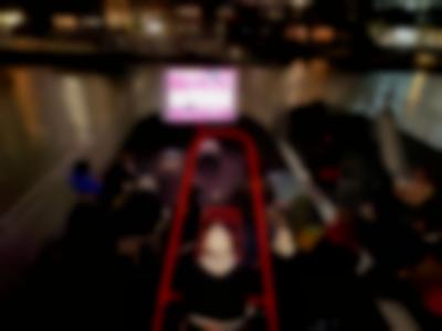 On a deck, viewed from above, a group of people stand or sit in front of a screen where something is being projected. To their right, there is a slide. In front of the screen, there is a red playground overhead ladder, behind which someone is standing, wearing headphones. In the background, behind the screen, there is a river and two large buildings on the other bank. It is dark and several of the windows in the buildings have lights on.
