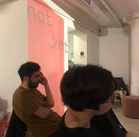 Two people sit facing the same direction. Behind them, there is wall projection of a red block with the words 
