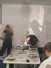 At the end of a long table, on top of which there are several sheets of paper, a person is leaning over an overhead projector. They are making notes on the sheet on the projector's stage. Next to them, someone stands in front of a flip chart easel.