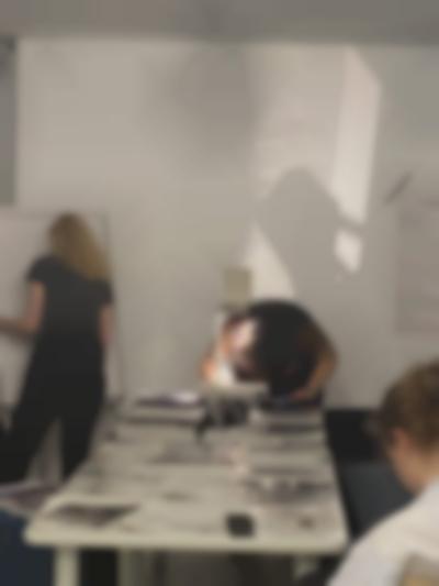 At the end of a long table, on top of which there are several sheets of paper, a person is leaning over an overhead projector. They are making notes on the sheet on the projector's stage. Next to them, someone stands in front of a flip chart easel.