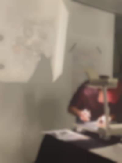 A person is leaning over an overhead projector. They are making notes on the sheet on the projector's stage. The image is being projected behind them. Next to the projection, a white sheet of paper with notes, handwritten in different colours, has been stuck to the wall with two thin strips of black tape.