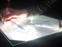 On top of a overhead project's stage, someone is making notes in red on a piece of glass. Underneath it, there is a printed text.