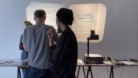 Four people stand around a table, on top of which there is an overhead projector and other materials. On the wall, two projections show documents with handwritten notes.
