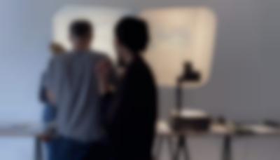 Four people stand around a table, on top of which there is an overhead projector and other materials. On the wall, two projections show documents with handwritten notes.