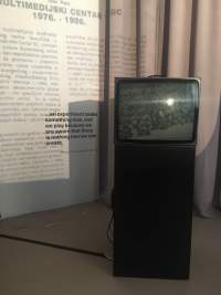 A television box has been set on a black plinth. Behind it, there are beige curtains a white wall, onto which a text is being projected.