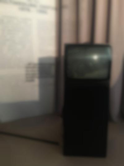 A television box has been set on a black plinth. Behind it, there are beige curtains a white wall, onto which a text is being projected.
