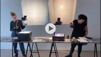Two people stand behind a long table, each next to one of the two overhead projectors. They both look at things on the table, on which several other printed materials have been laid. The projectors are shining a light behind the two people, but no image is being projected. At the centre of the image, there is a dark grey triangle inside a grey circle, to indicate a 