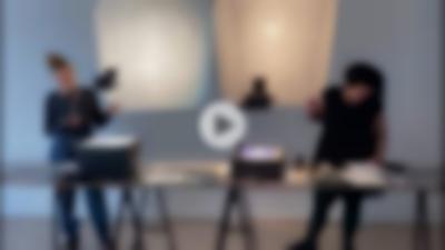 Two people stand behind a long table, each next to one of the two overhead projectors. They both look at things on the table, on which several other printed materials have been laid. The projectors are shining a light behind the two people, but no image is being projected. At the centre of the image, there is a dark grey triangle inside a grey circle, to indicate a 