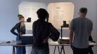 Four people stand around a table, on top of which there are two overhead projectors and other materials. On the wall, two projections show documents with handwritten notes.