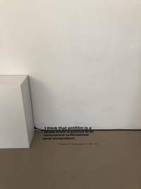 A wall text appears at the bottom of a wall; the first line appears on the wall, with the remaining of the text on the beige floor. To the left, a white plinth remains empty. The text reads 
