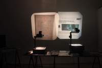 In a dark room, two overhead projectors project two images. On the left, an old document on beige paper. On the right, a blue photograph with a close-up of a person's face and some text in black around it.