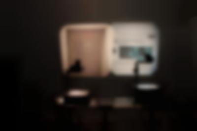 In a dark room, two overhead projectors project two images. On the left, an old document on beige paper. On the right, a blue photograph with a close-up of a person's face and some text in black around it.