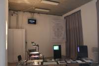 Small indoor space with large beige curtain. Next to the curtain, there is a large white textile with a printed diagram hanging from near the ceiling. On each side of the curtains, there is one television box on a plinth. Across, two overhead projectors are projecting on the opposite wall. At the front, there is a table with other printed materials.