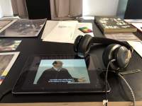 Table seen from above. On the table, there is a tablet on which the image of a masculine-presenting person gesticulating with their left hand can be seen. There are subtitles on the image. A set of headphones has been connected to the device, and lays on the table next to it. Other books and printed material can also be seen on the table.