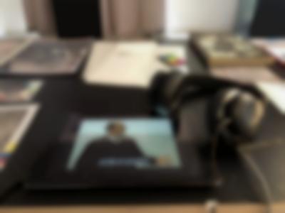 Table seen from above. On the table, there is a tablet on which the image of a masculine-presenting person gesticulating with their left hand can be seen. There are subtitles on the image. A set of headphones has been connected to the device, and lays on the table next to it. Other books and printed material can also be seen on the table.