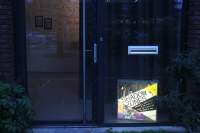 Ground level view of building front, with a glass door and a glass floor to ceiling window with letterbox. On the floor, window side, there is a square light box, in which 