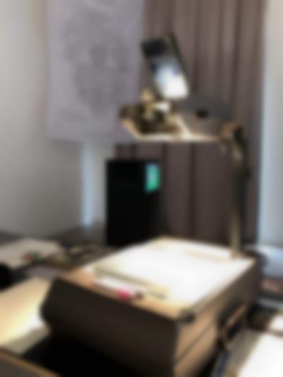 Overhead projector, shining a strong light towards its stage. Behind it, there is large beige curtain and a white textile with a printed diagram hanging from near the ceiling. In front of the curtain, there is one television box on a plinth.