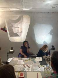 Four people sit around a table, on top of which there is printed material and pens and pencils. There two overhead projectors, one on each side of the table, projecting images to a wall. One of the people is writing on a sheet of paper, whose image is being projected on the wall.
