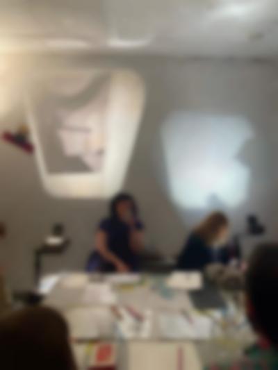 Four people sit around a table, on top of which there is printed material and pens and pencils. There two overhead projectors, one on each side of the table, projecting images to a wall. One of the people is writing on a sheet of paper, whose image is being projected on the wall.
