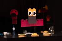 Hanging installation, made from legos: at the bottom, a long red lego has been left horizontal. A brown and yellow lego with two eye cartoon stickers has been placed vertically slightly higher, in front. Behind it, other hanging structures are discernible, as well as a table with a few plates with food.