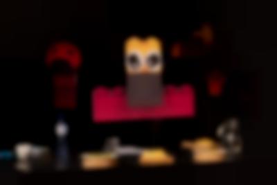 Hanging installation, made from legos: at the bottom, a long red lego has been left horizontal. A brown and yellow lego with two eye cartoon stickers has been placed vertically slightly higher, in front. Behind it, other hanging structures are discernible, as well as a table with a few plates with food.