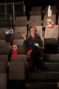 A person sits in tribute seating, with their left leg crossed. They are wearing headphones and holding a white cup. Around them, there are several installations hanging from above, mostly made from legos. Two of them have small cartoon eyes attached.