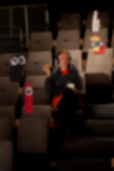 A person sits in tribute seating, with their left leg crossed. They are wearing headphones and holding a white cup. Around them, there are several installations hanging from above, mostly made from legos. Two of them have small cartoon eyes attached.